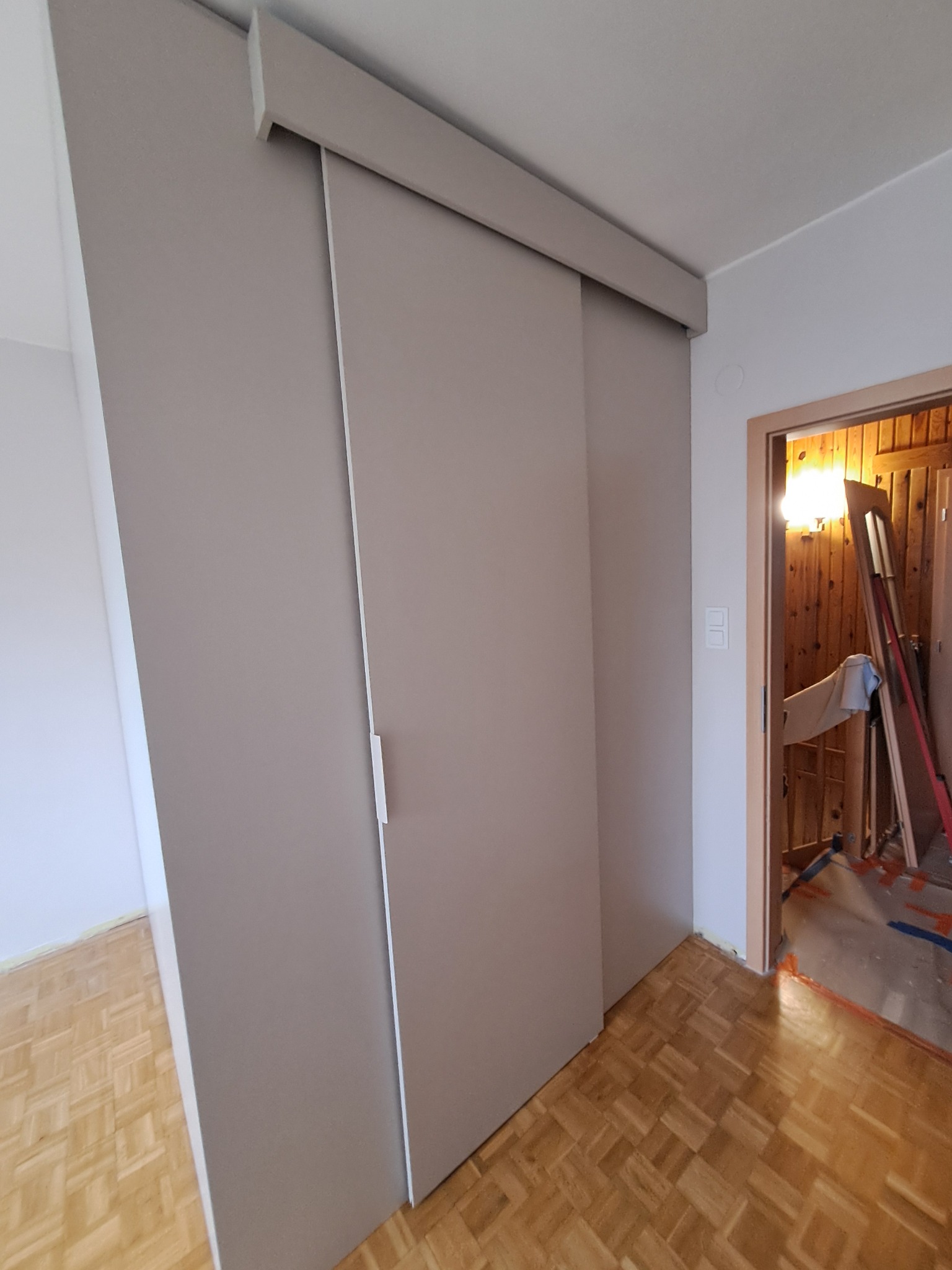 Zamknięta szafa przesuwna z gładkimi, beżowymi drzwiami w jasnym pokoju, widoczny fragment parkietu i wnętrze innego pomieszczenia w trakcie remontu z drabiną i narzędziami.