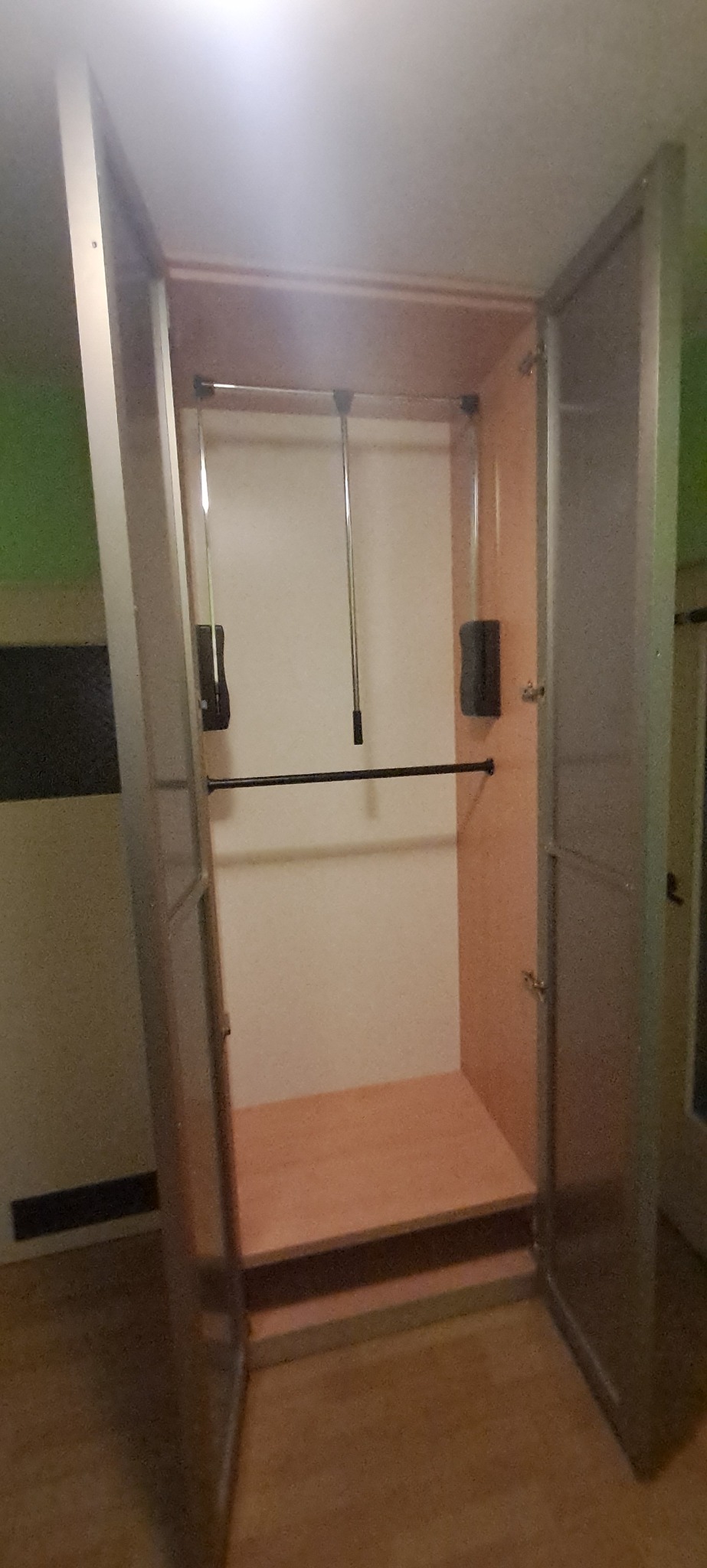 Wnętrze otwartej szafy z półką i drążkami na ubrania, widoczne mechanizmy do opuszczania drążków, jasne wnętrze szafy, szare drzwi.