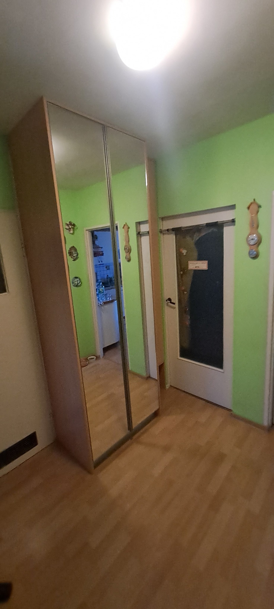 Szafa wnękowa z lustrzanymi drzwiami przesuwnymi, odbijająca wnętrze mieszkania z zielonymi ścianami i drewnianą podłogą.