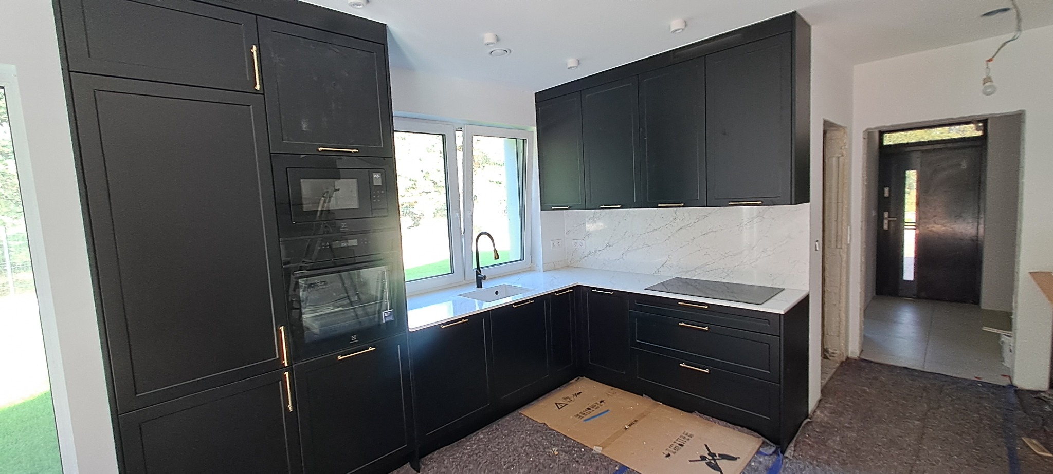 Nowoczesna kuchnia z czarnymi szafkami z frezowanymi frontami i złotymi uchwytami, biały blat z marmurowym wzorem, zabudowany piekarnik i mikrofalówka, widoczny karton na podłodze sugerujący prace...
