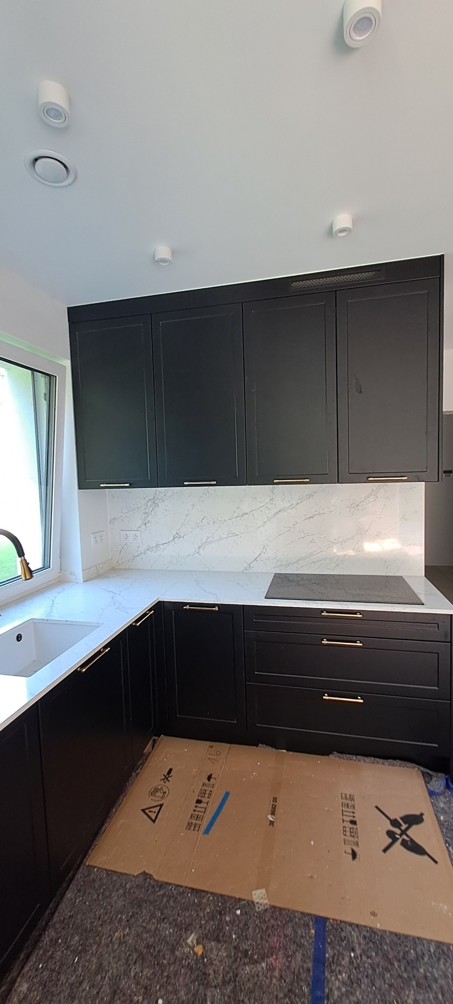 Czarne szafki kuchenne z złotymi uchwytami i blatem imitującym marmur, chronione kartonem na podłodze podczas montażu.