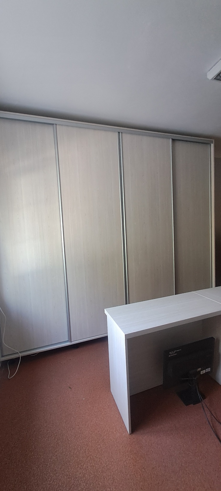 Beżowa szafa wnękowa z przesuwnymi drzwiami i srebrnymi obramowaniami, stojąca obok jasnego biurka z widocznym monitorem, na tle pomarańczowej podłogi.