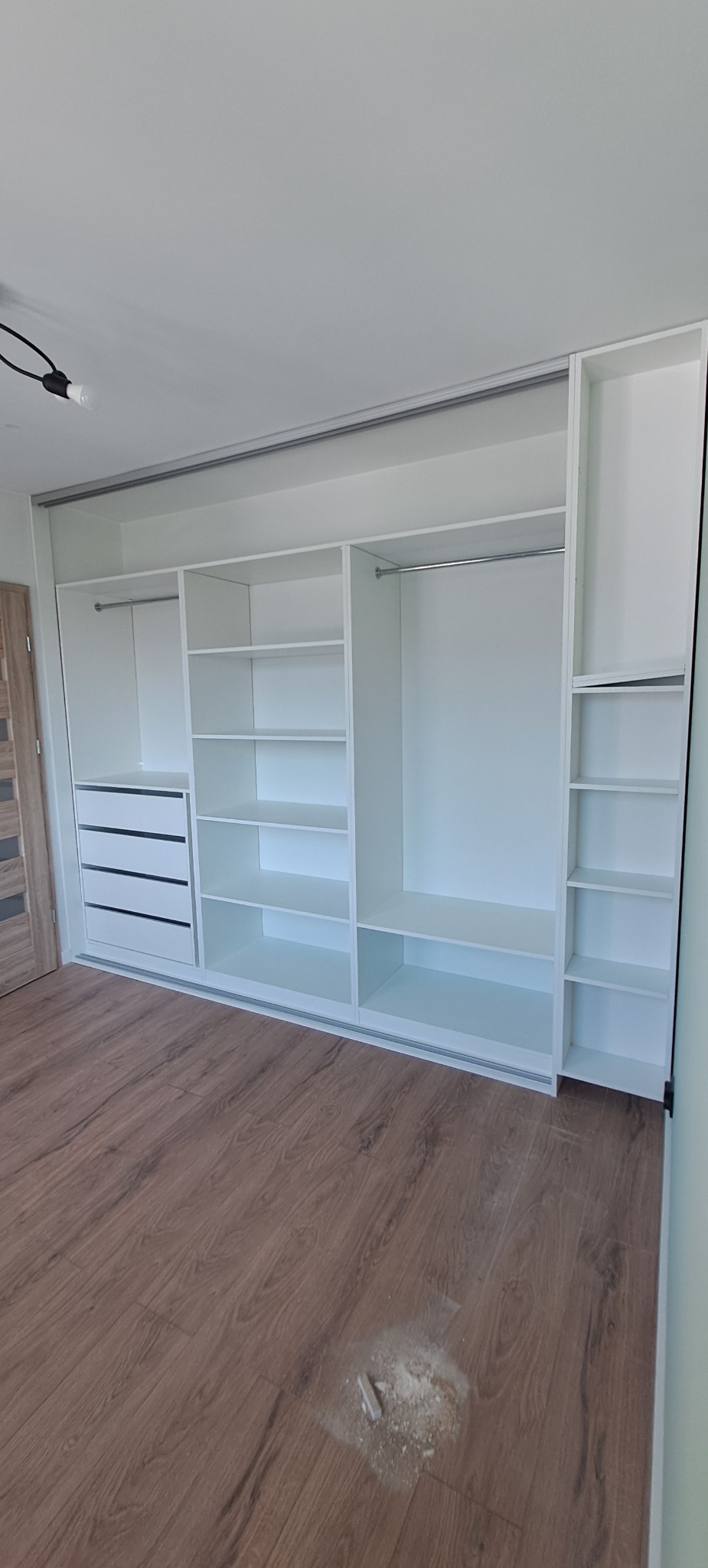 Biała szafa wnękowa z przesuwnymi drzwiami, z półkami i szufladami, na podłodze widoczne resztki po montażu.