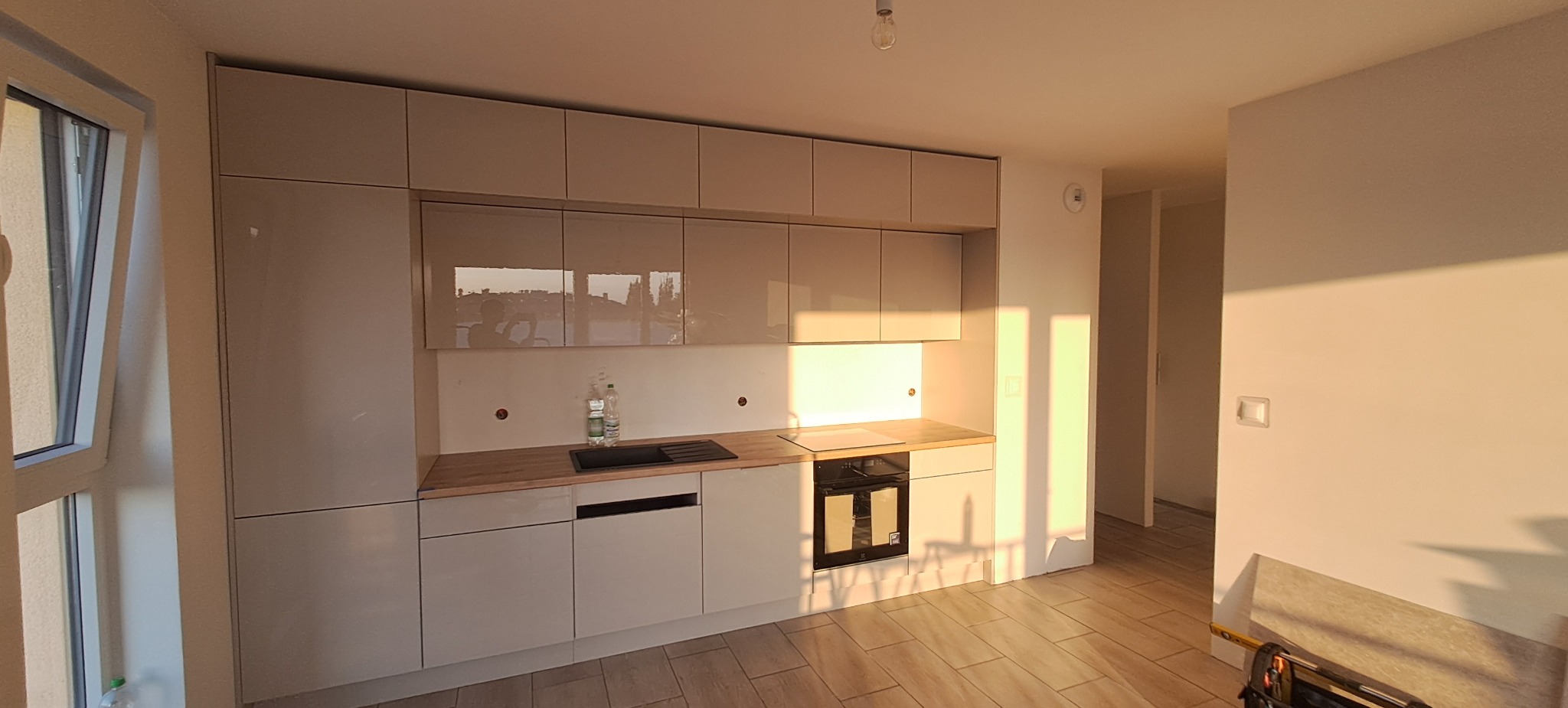 Jasna, nowoczesna kuchnia z błyszczącymi frontami, drewnianym blatem i czarnym zlewem, oświetlona popołudniowym słońcem w nowym mieszkaniu, widoczne niedokończone wykończenia i narzędzia.