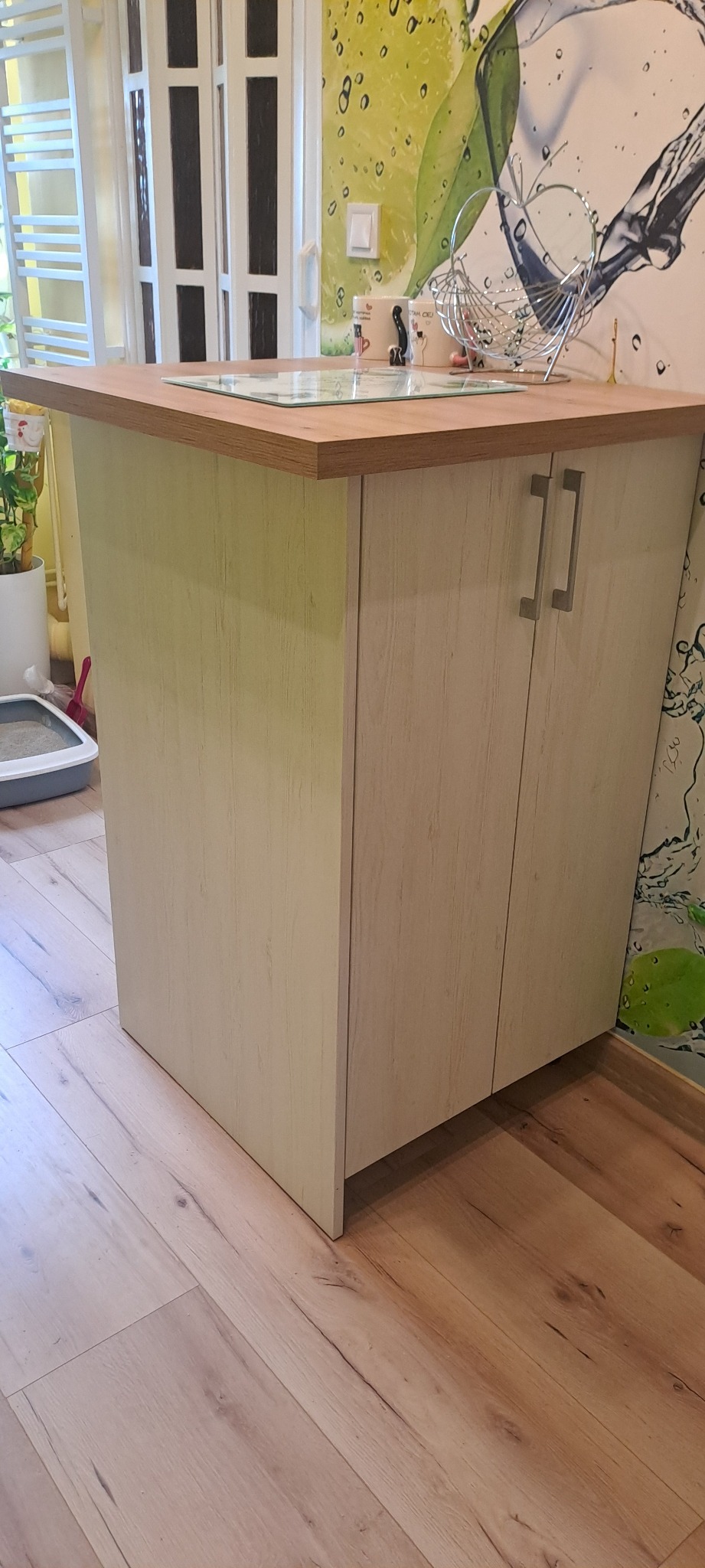 Wykończona szafka kuchenna z blatem z drewna, jasne fronty szafek z podłużnymi uchwytami, na blacie szklana płyta, w tle wzorzysta tapeta z motywem owoców i wody.