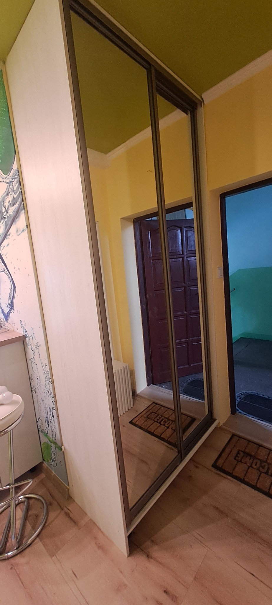 Biała szafa wnękowa z przesuwnymi drzwiami lustrzanymi, widoczna w korytarzu z żółtymi ścianami i zielonym sufitem, z odblitym w lustrze fragmentem innego pokoju.