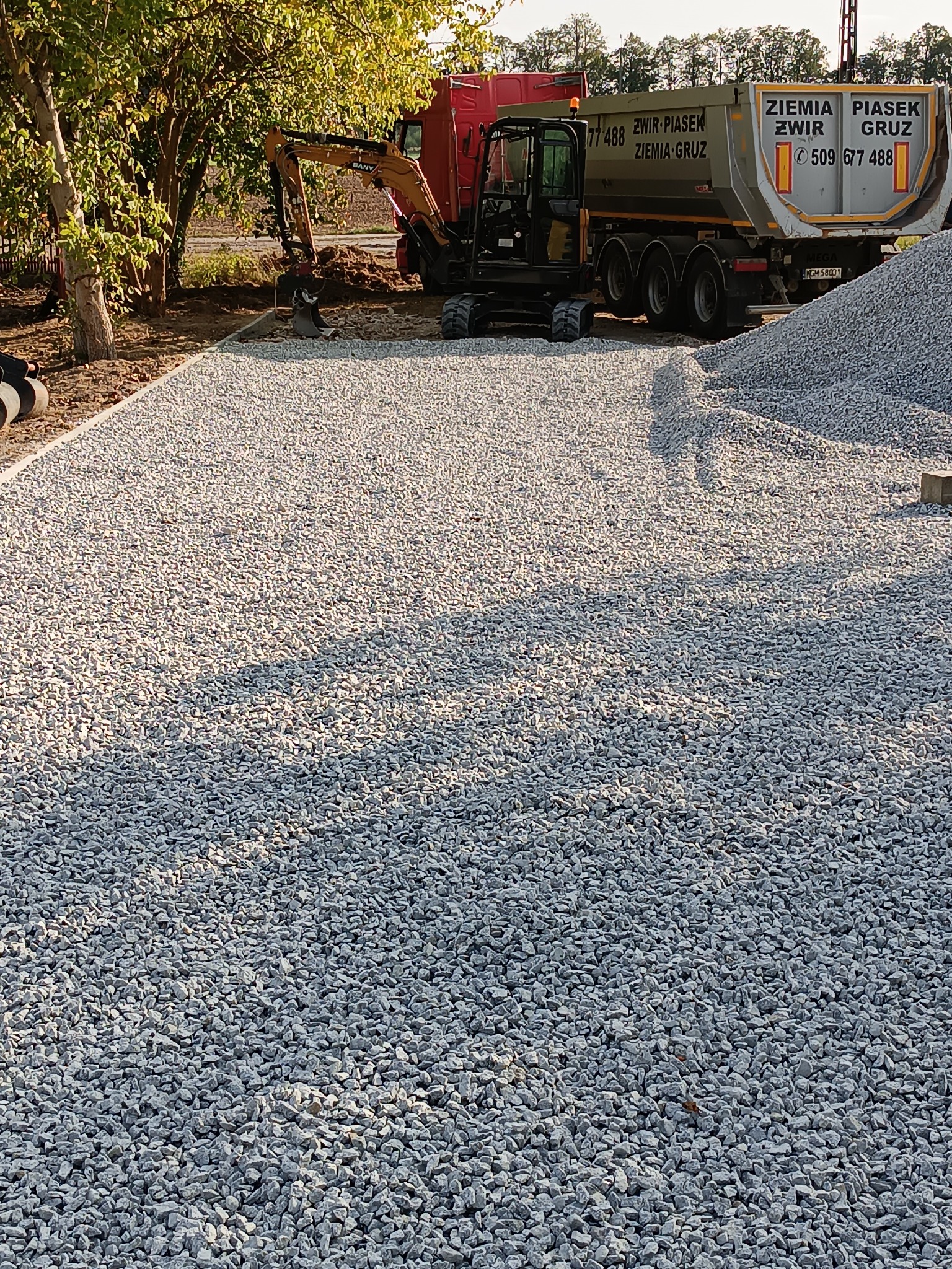 Obszar pokryty drobnym, szarym kruszywem, z widocznym w tle czerwonym samochodem ciężarowym z napisem 'Ziemia Piasek Żwir Gruz' oraz małą koparką, sugerujący przygotowanie terenu pod budowę...