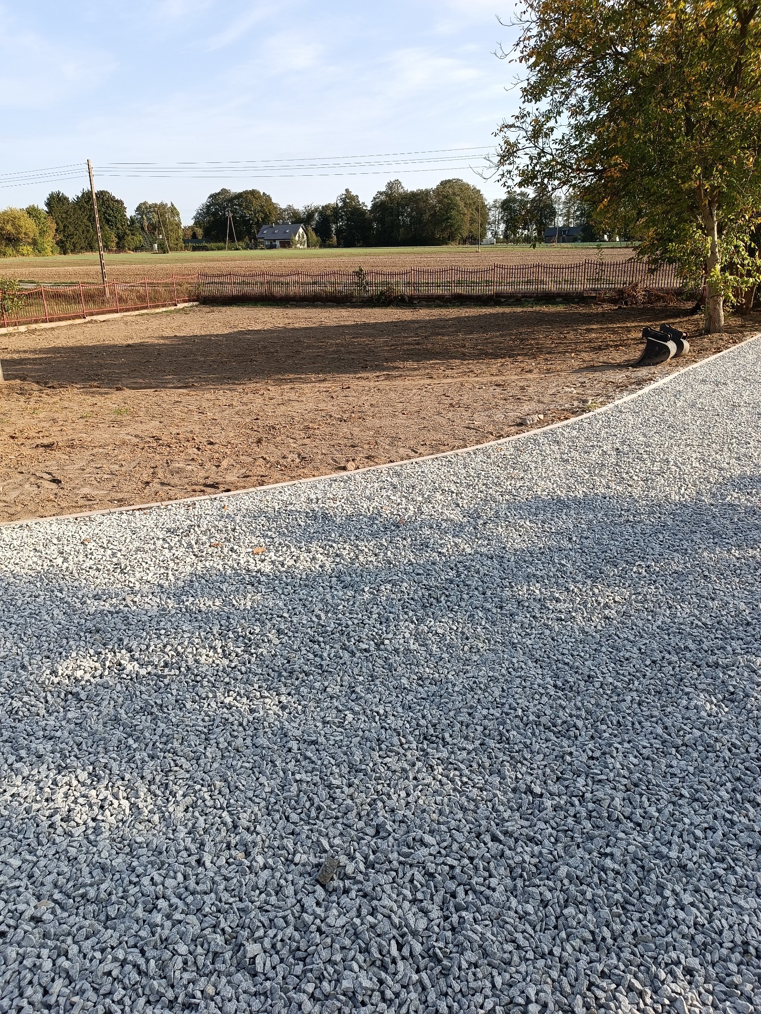 Wykończona ścieżka z jasnoszarego kruszywa oddzielona obrzeżem od przygotowanego podłoża pod trawnik, w tle widoczny krajobraz z drzewami i domem.