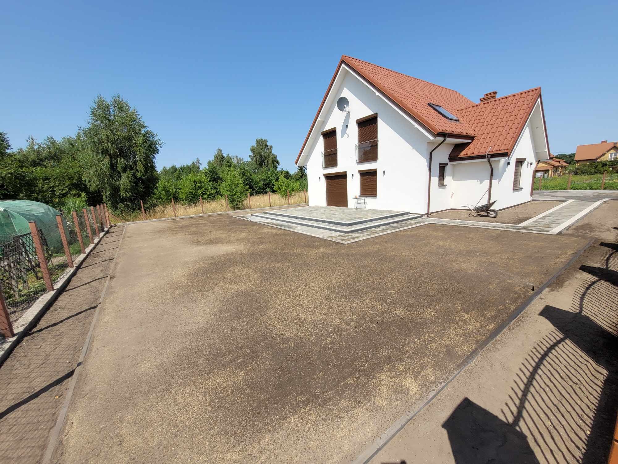 Przygotowany teren pod trawnik z widocznym siewem nasion trawy, nowy dom z czerwoną dachówką w tle, obrzeża z kostki betonowej i częściowo wykonany chodnik z kostki. W tle drzewa i błękitne niebo.