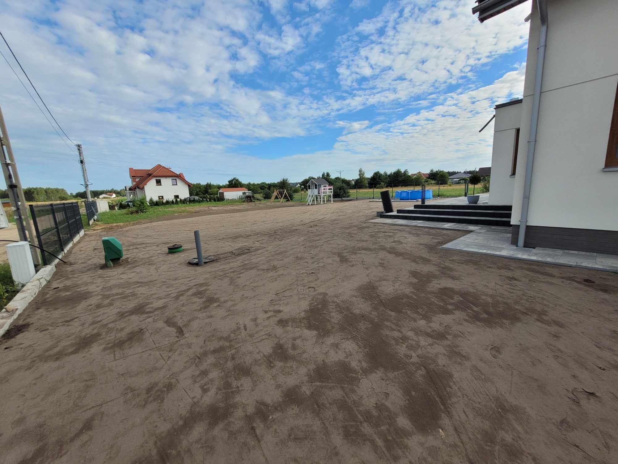 Przygotowany teren pod trawnik z widocznymi śladami po maszynach, w tle dom z czerwonym dachem, plac zabaw i basen.