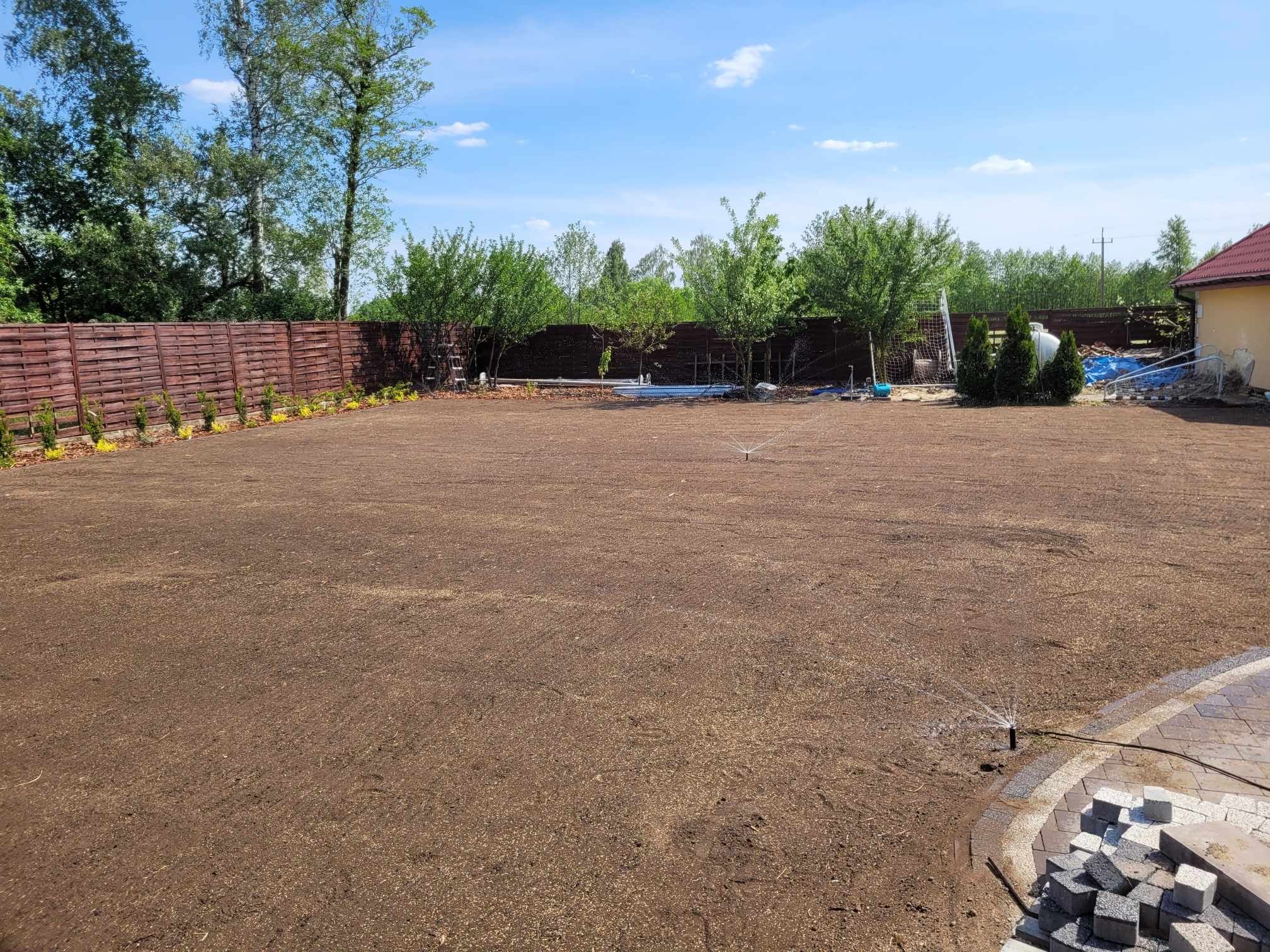 Przygotowany teren pod trawnik z widocznymi zraszaczami nawadniającymi glebę, obok ułożone kostki brukowe, w tle ogrodzenie z drewnianych paneli i zieleń drzew.