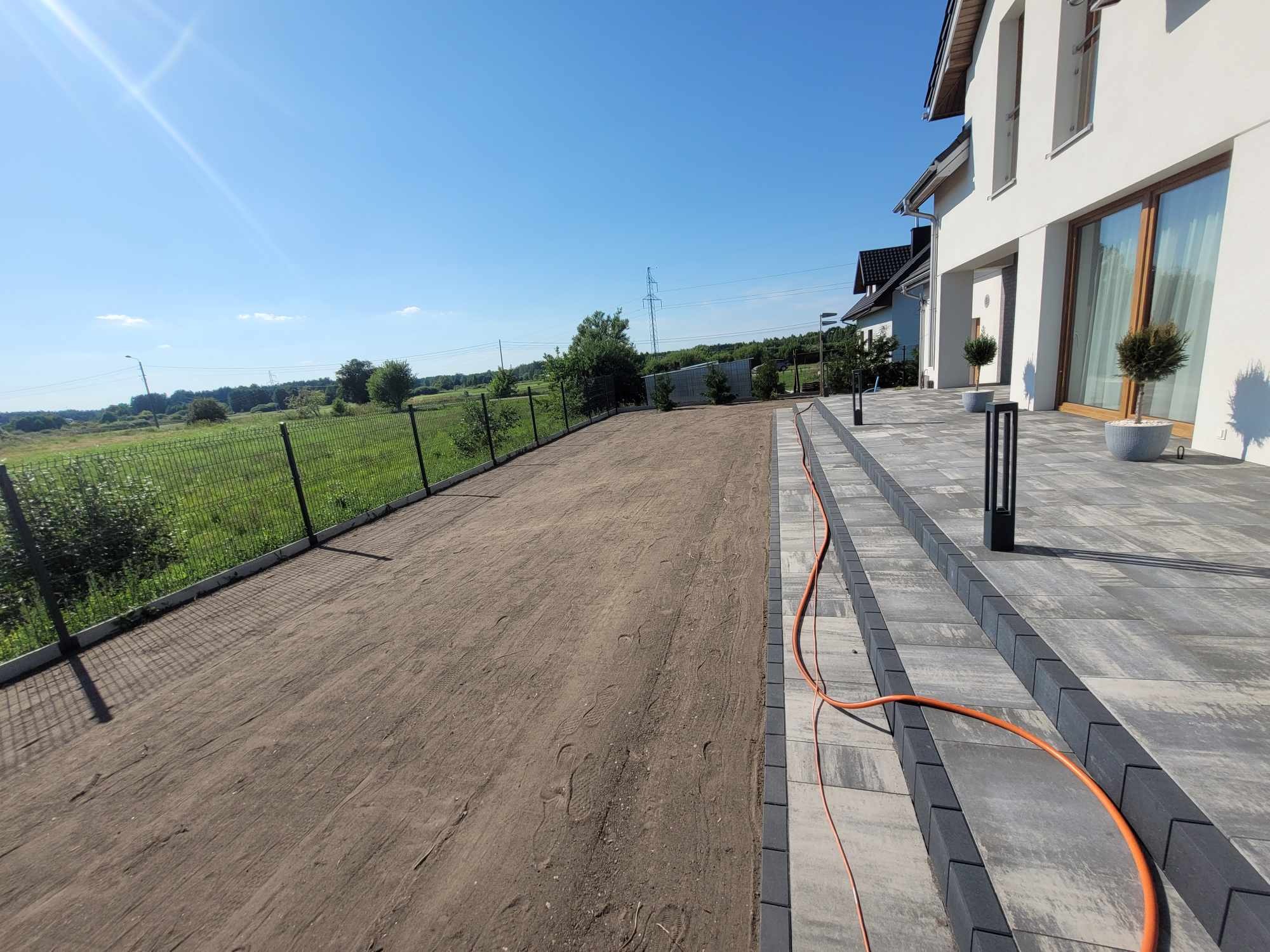 Przygotowany teren pod trawnik z rolki, widoczne ślady stóp, obrzeża z kostki, nowoczesny dom z szarym tarasem i lampami ogrodowymi, pomarańczowy przewód na tarasie, zielone pole za ogrodzeniem.