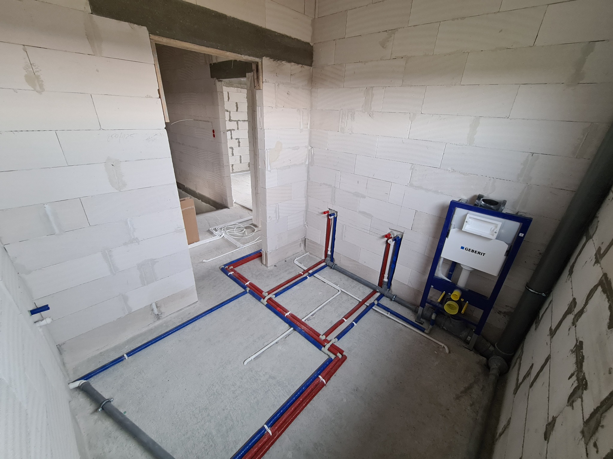 Surowy stan łazienki z widoczną instalacją wodną z rur czerwonych i niebieskich oraz stelażem podtynkowym Geberit, ściany z białego betonu komórkowego.