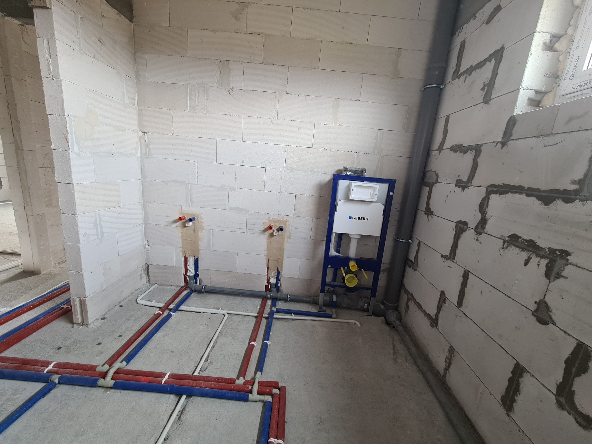 Surowy stan łazienki z widoczną instalacją podtynkową Geberit, rurami ciepłej i zimnej wody w czerwono-niebieskiej izolacji oraz szarą rurą kanalizacyjną przy ścianie z betonu komórkowego.