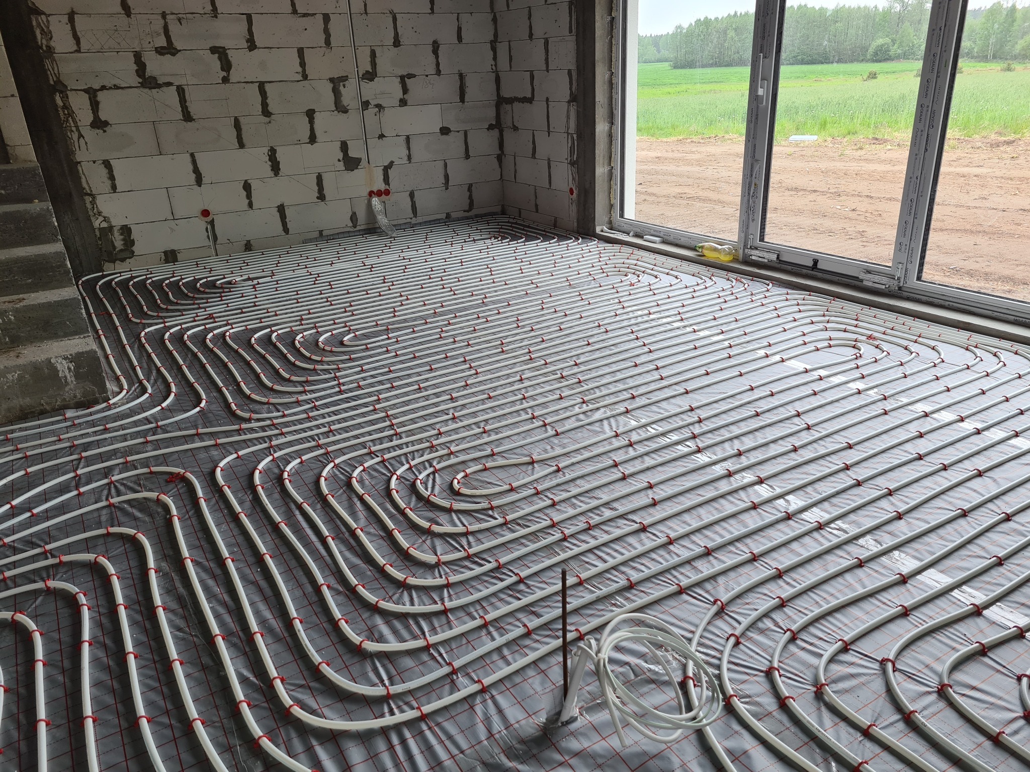 Układanie rur ogrzewania podłogowego na folii z siatką w surowym wnętrzu domu w trakcie budowy, widok na zielone pole za oknem.