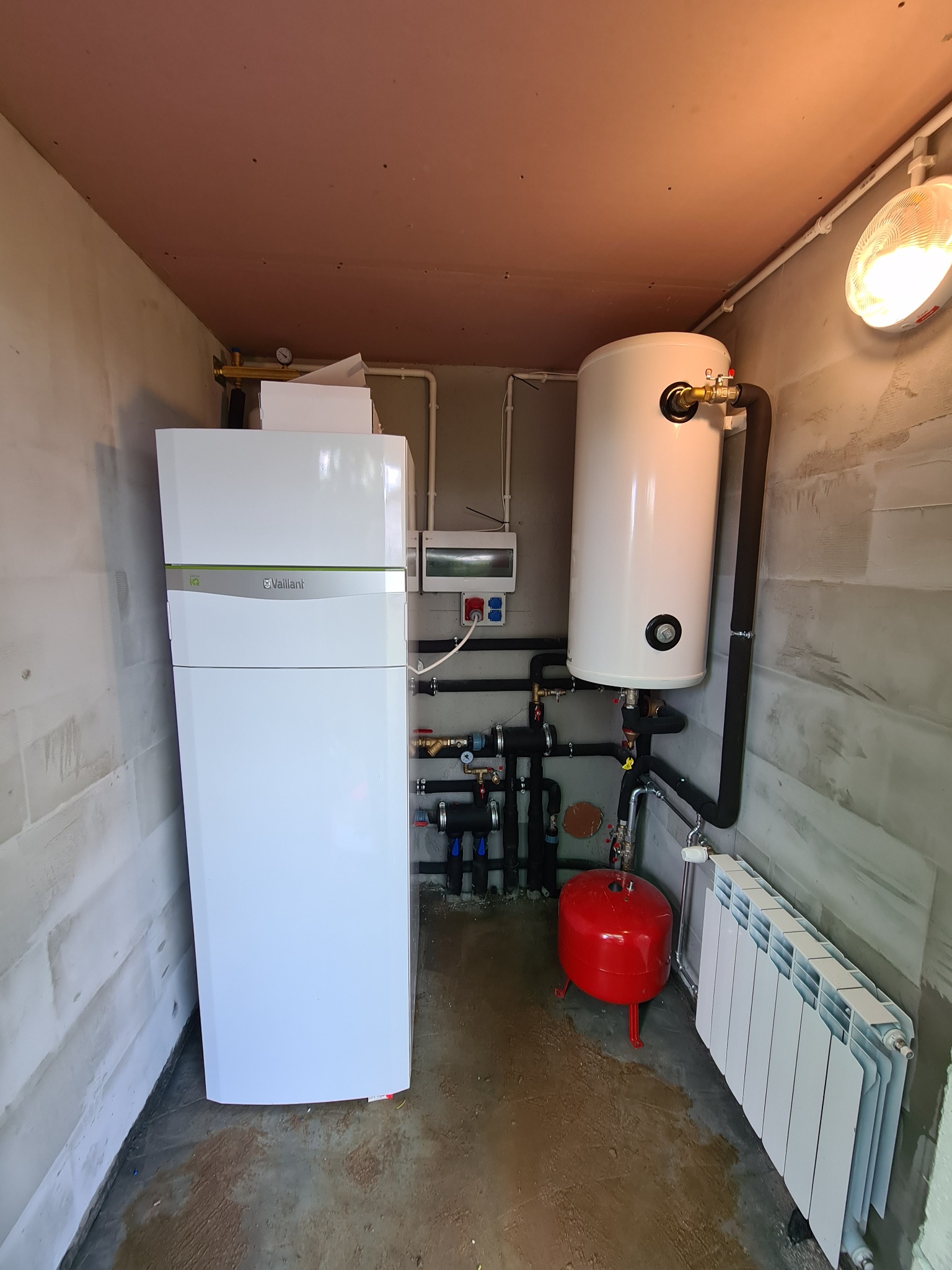 Zestaw urządzeń grzewczych w pomieszczeniu technicznym w Biskupcu: biały piec gazowy Vaillant, biały zasobnik ciepłej wody, czerwone naczynie przeponowe i biały grzejnik panelowy.