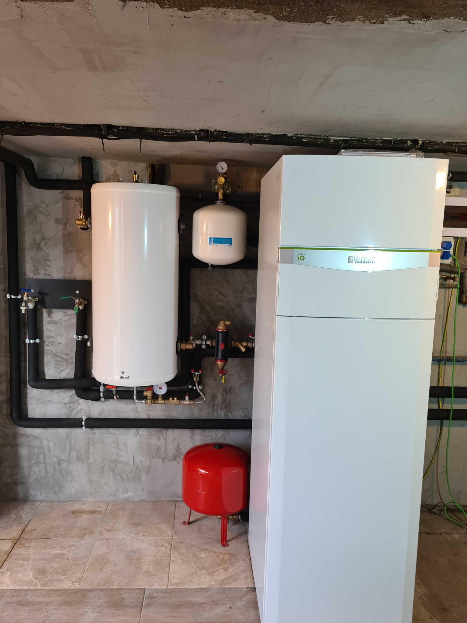 Biały zasobnik ciepłej wody użytkowej z czarnymi rurami i manometrami, obok biała pompa ciepła Vaillant z zielonym akcentem, czerwony zbiornik wyrównawczy na podłodze z płytek.