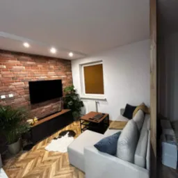 Salon z ceglaną ścianą, drewnianą podłogą w jodełkę i szarą sofą narożną; widoczne rośliny doniczkowe, stolik kawowy i telewizor.