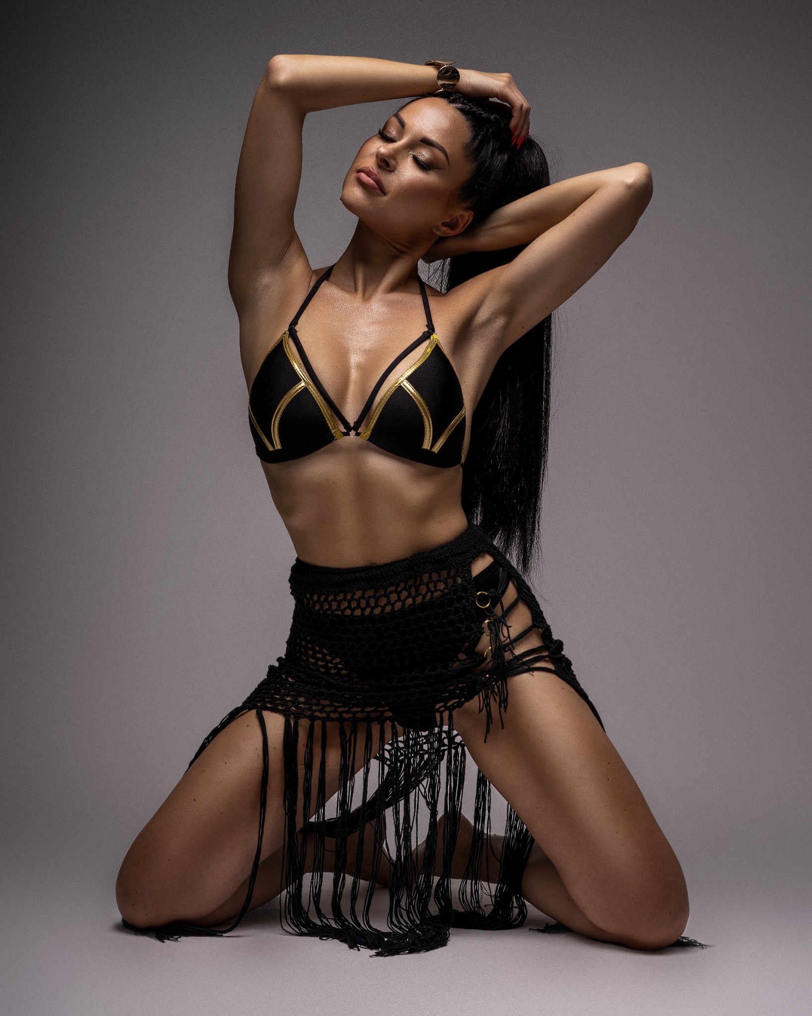 Kobieta w czarnym bikini z złotymi detalami i ażurowej spódnicy z frędzlami, pozuje na kolanach z zamkniętymi oczami i rękami uniesionymi nad głową, na jednolitym szarym tle.