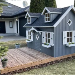 Szary drewniany domek dla dzieci z białymi okiennicami i doniczkami z kwiatami, umieszczony na drewnianym podeście w ogrodzie.