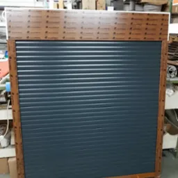 Szara, zwinięta roleta zewnętrzna z brązową ramą Alufrof, prezentowana w warsztacie lub magazynie.