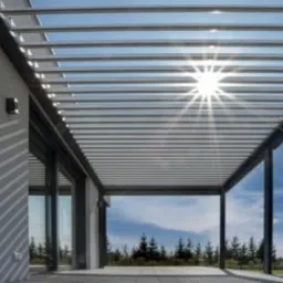 Nowoczesna pergola aluminiowa z poziomymi lamelami, przez które prześwieca słońce, widok z tarasu na ogród z drzewami i błękitnym niebem.