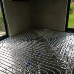 Szare pomieszczenie w trakcie montażu systemu rur na folii aluminiowej, przygotowanego do wylewki pod ogrzewanie podłogowe, widok z g&oacute;ry na ułożone pętle, z oknami z widokiem na zieleń w tle.