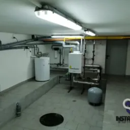 Kotłownia z białym bojlerem, piecem gazowym i szarymi rurami pod sufitem, oświetlona jarzeniówkami, z logo firmy instalacyjnej w prawym dolnym rogu.