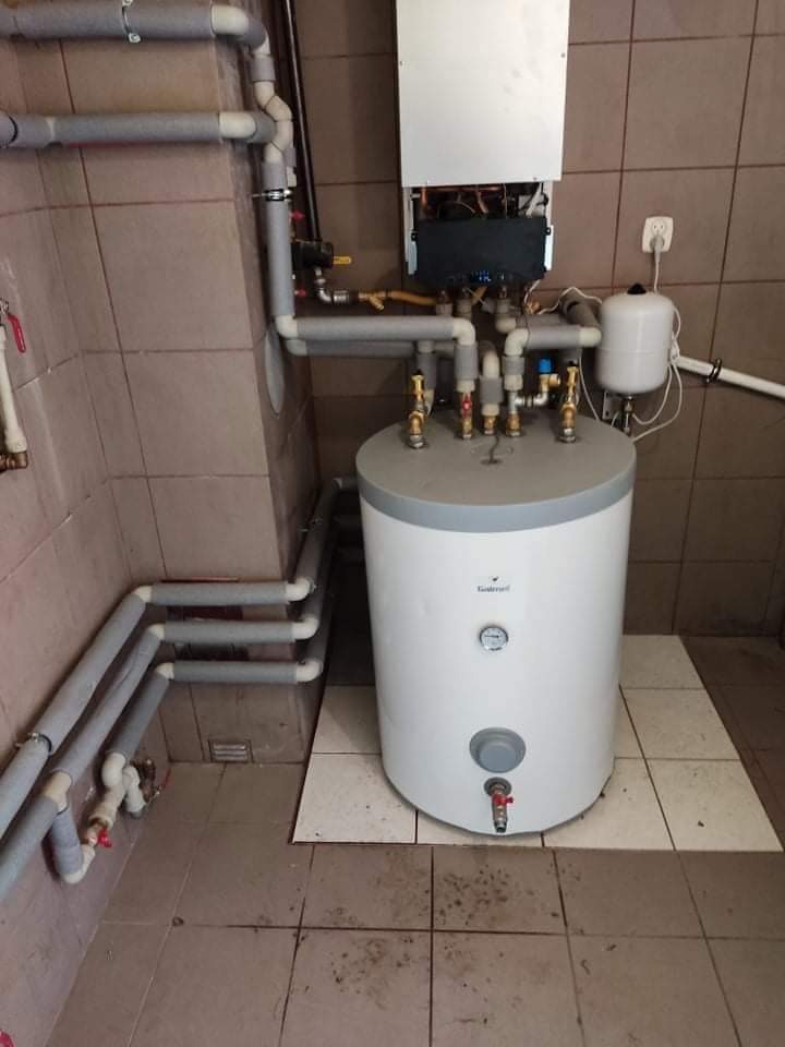 Bojler z podłączonymi rurami w kotłowni wyłożonej kafelkami, widoczny również piec gazowy na ścianie.