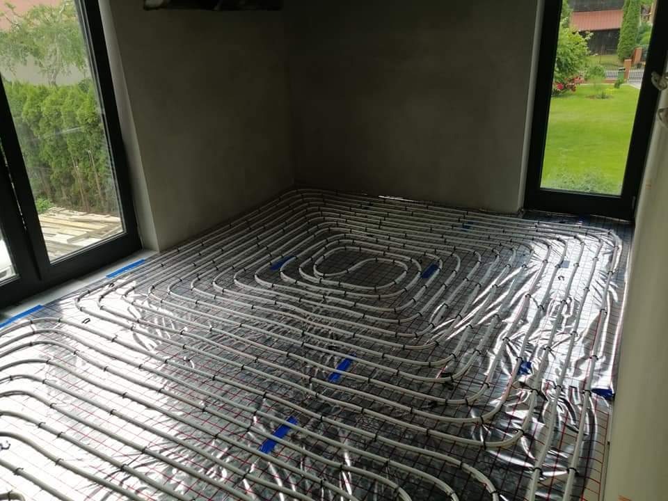 Szare pomieszczenie w trakcie montażu systemu rur na folii aluminiowej, przygotowanego do wylewki pod ogrzewanie podłogowe, widok z góry na ułożone pętle, z oknami z widokiem na zieleń w tle.