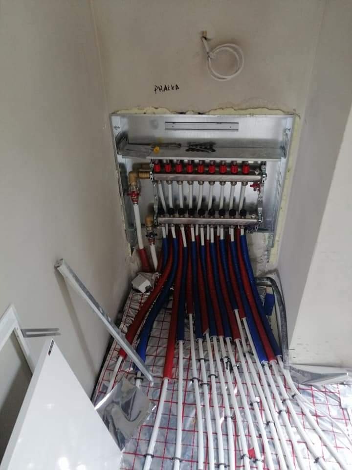 Rozdzielacz instalacji grzewczej podłogowej z rurami w kolorach czerwonym i niebieskim, ułożonymi na folii izolacyjnej z nadrukowaną siatką, widoczne niedokończone wykończenie ściany i sufitu.