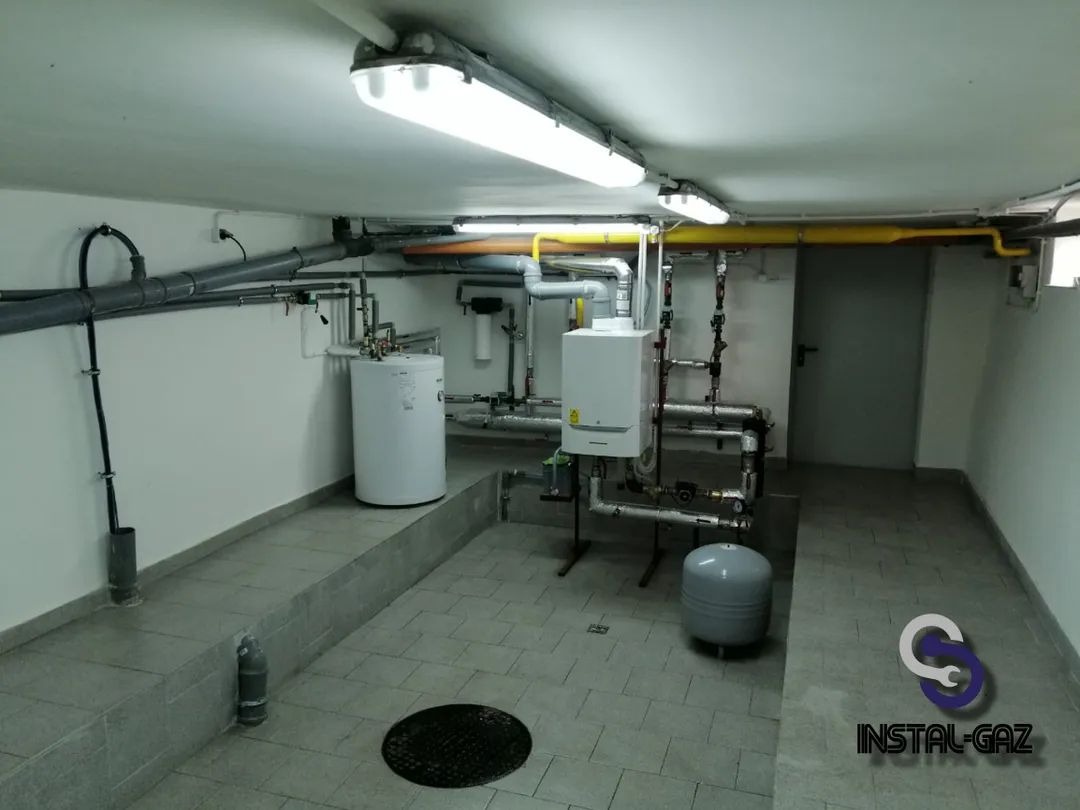 Kotłownia z białym bojlerem, piecem gazowym i szarymi rurami pod sufitem, oświetlona jarzeniówkami, z logo firmy instalacyjnej w prawym dolnym rogu.