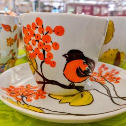 Herbaty i Kawy Świata - Biała filiżanka z motywem jarzębiny i ptaszka siedząca na dopasowanym talerzyku, obok widoczne inne filiżanki z motywami jesiennymi, całość na zielonym tle.