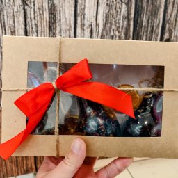 Herbaty i Kawy Świata - Ręka trzyma kartonowe pudełko prezentowe z okienkiem, przez które widać dekoracyjne świece i suszone pomarańcze, przewiązane sznurkiem jutowym i czerwoną wstążką na tle drewnianej ściany.