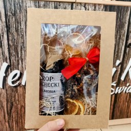 Herbaty i Kawy Świata - Prezent w kartonowym pudełku z okienkiem, zawierający butelkę syropu z aronii, suszone plastry pomarańczy i mieszankę herbaty, przewiązany czerwoną wstążką, na tle kartonowego opakowania...