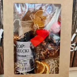 Ręcznie wykonany kosz prezentowy z dżemem z aronii, suszonymi pomarańczami i herbatą, zapakowany w tekturowe pudełko z okienkiem, przewiązany czerwoną wstążką.