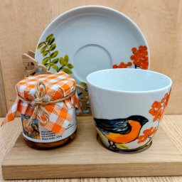 Herbaty i Kawy Świata - Słoik z dżemem pigwowym, nakryty pomarańczowo-białą kratką i sznurkiem, obok filiżanka z ręcznie malowanym ptakiem i jarzębiną oraz talerzyk z motywem roślinnym, na drewnianej desce.