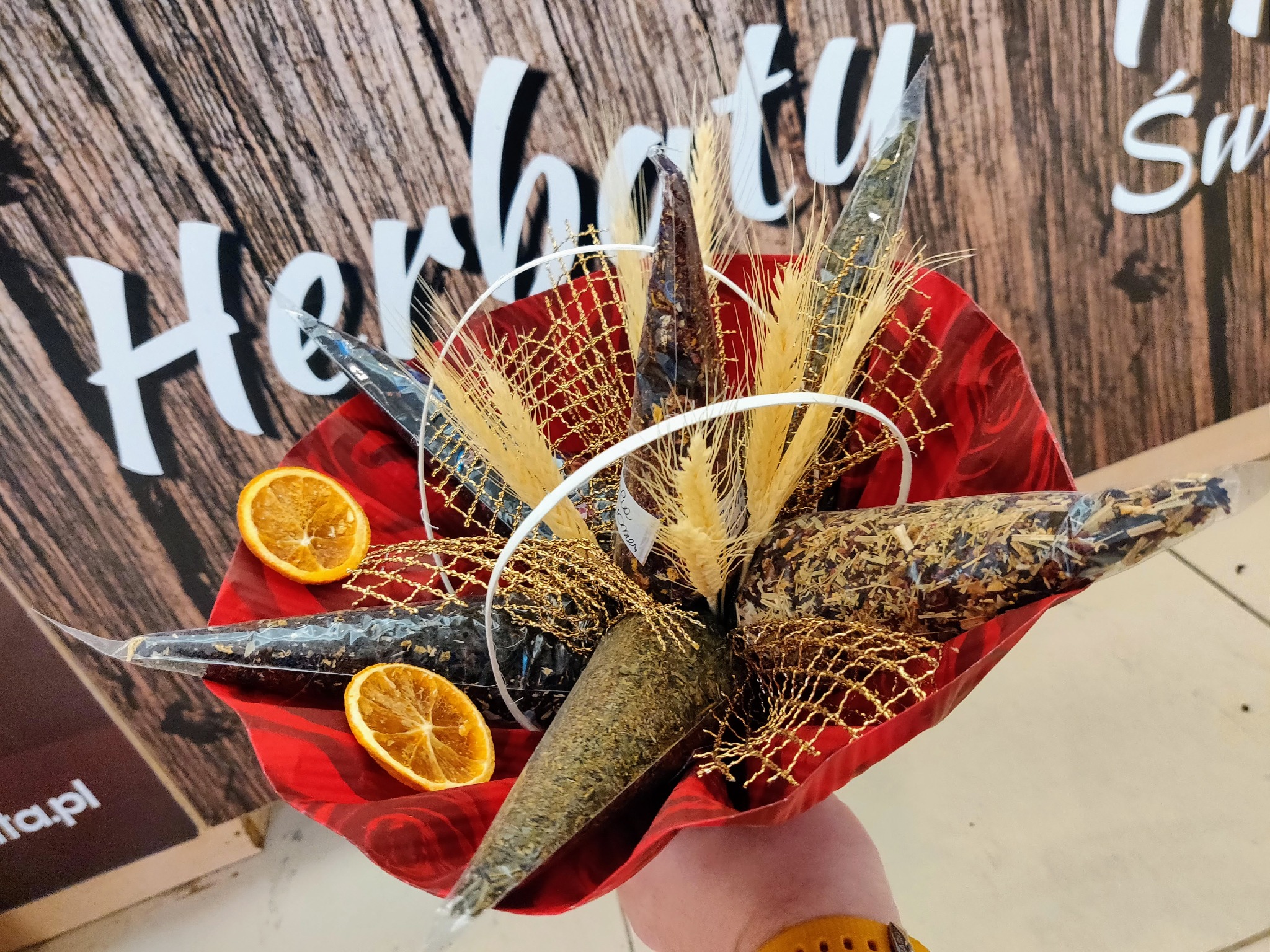 Ręcznie robiony bukiet z herbat w stożkowych opakowaniach, ozdobiony suszonymi pomarańczami, złotą siatką i źdźbłami zboża, zapakowany w czerwoną bibułę na tle drewnianej ściany z napisem.