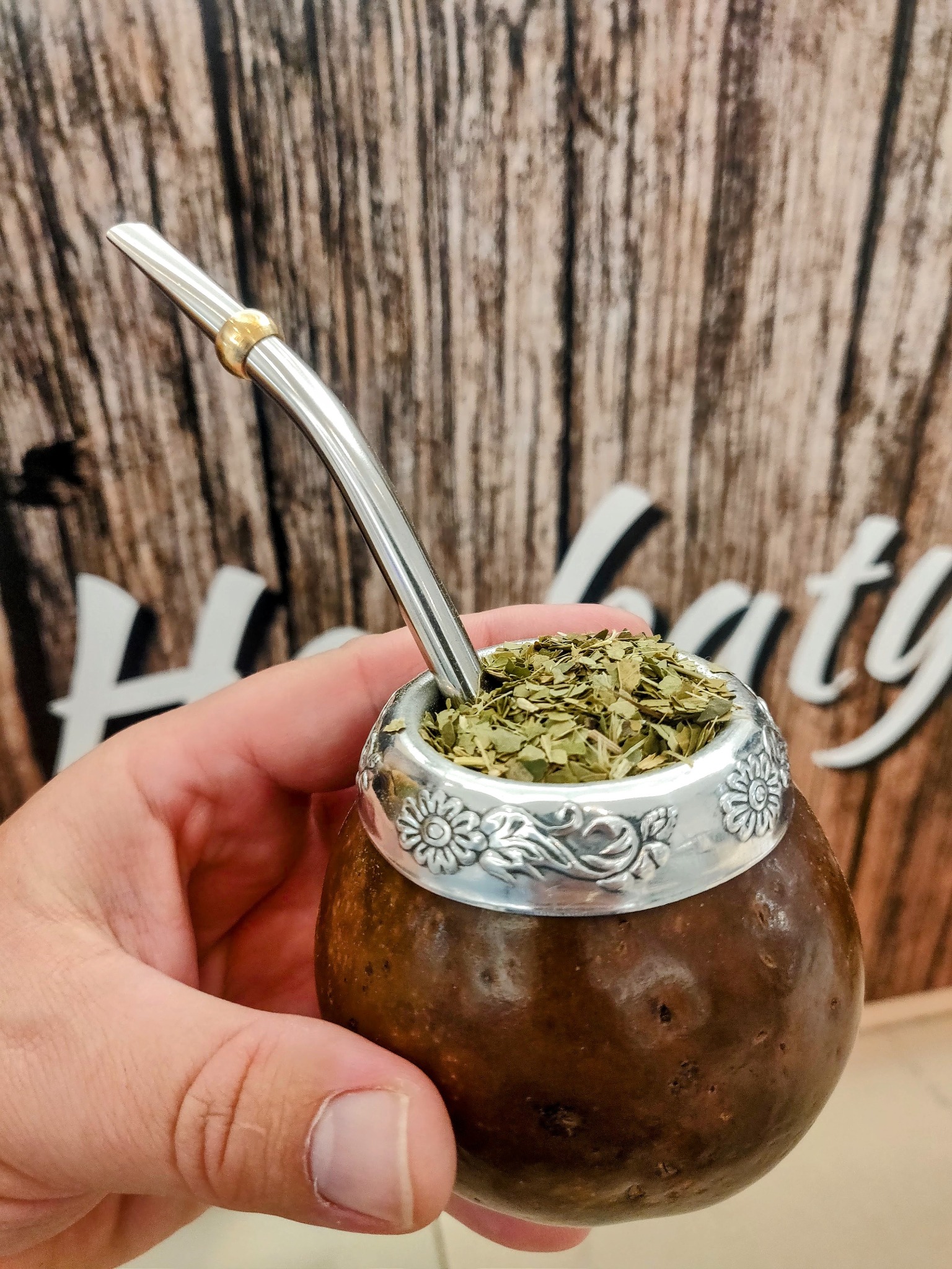 Ręka trzyma naczynie matero z yerba mate i bombillą na tle drewnianej ściany.