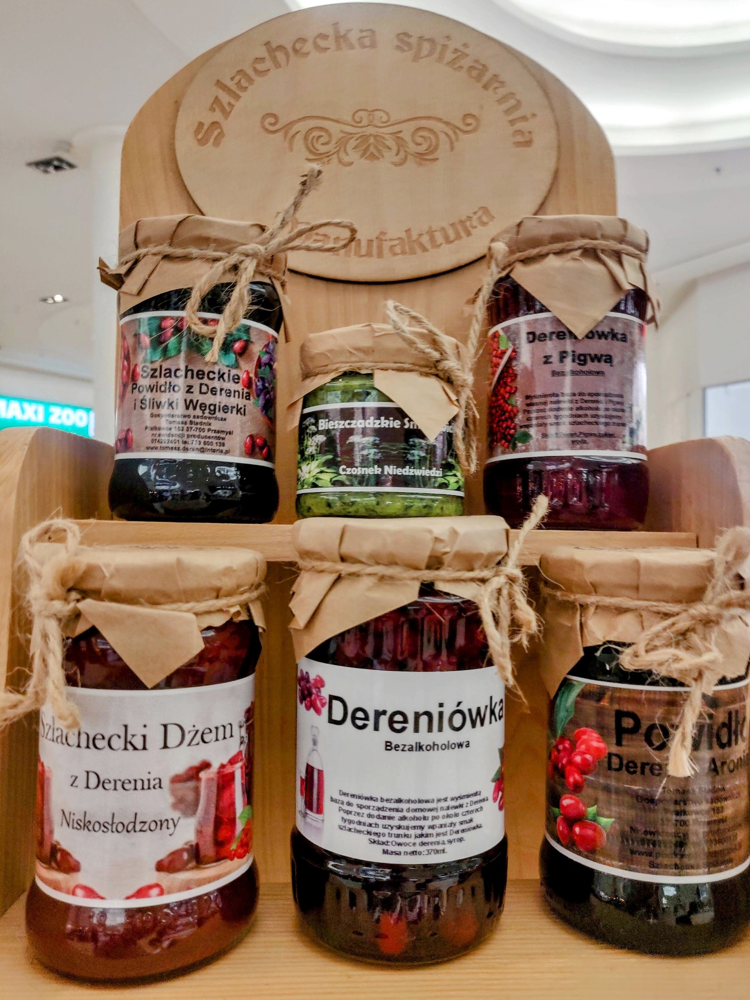 Półka z sześcioma słoikami przetworów owocowych i warzywnych, z etykietami w języku polskim, na drewnianym regale z wygrawerowanym napisem 'Szlachecka Spiżarnia Manufaktura'.