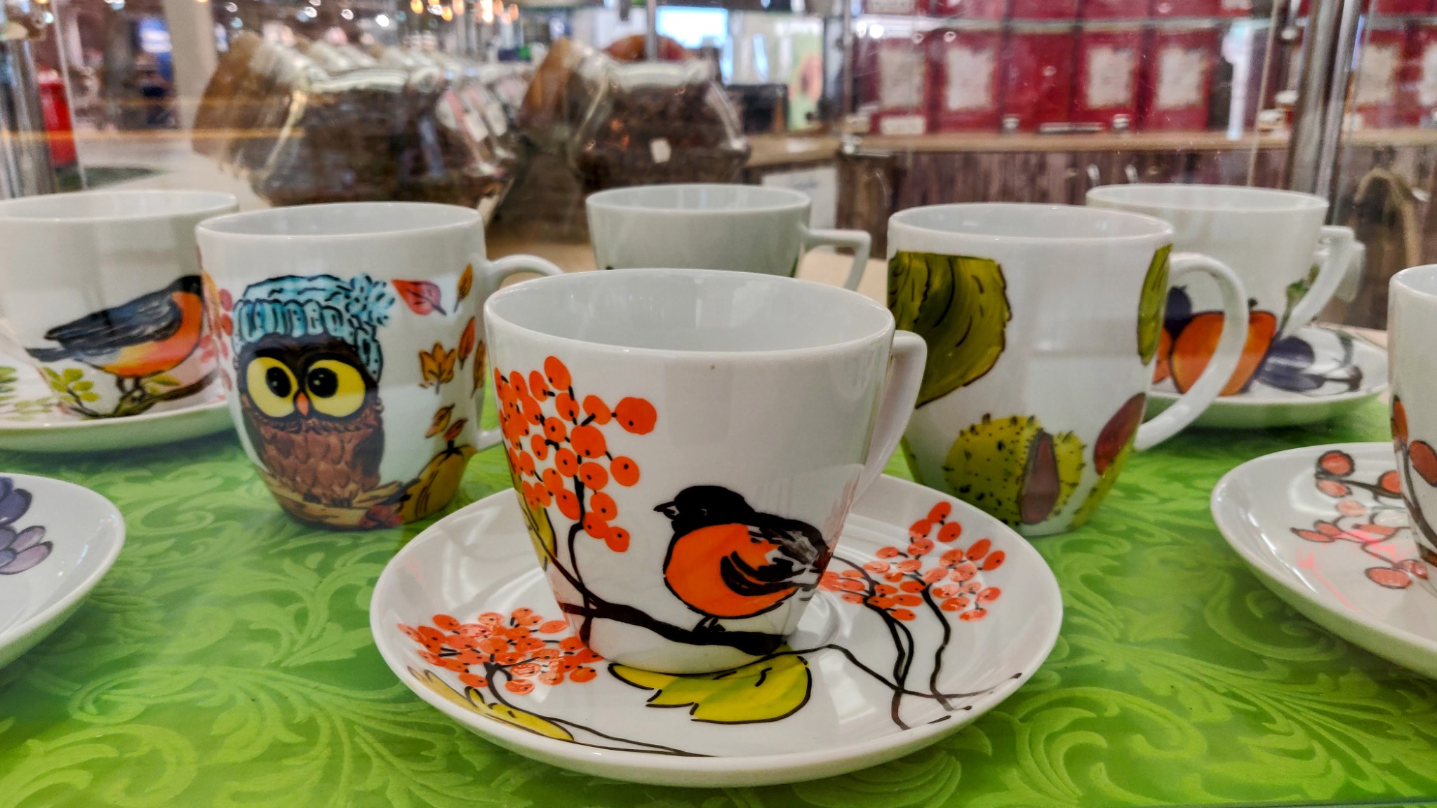 Rząd białych filiżanek i spodków z różnymi motywami zwierzęcymi i roślinnymi, w tym ptaków, sowy w czapce i owoców jarzębiny, ustawionych na zielonym obrusie ze wzorem.