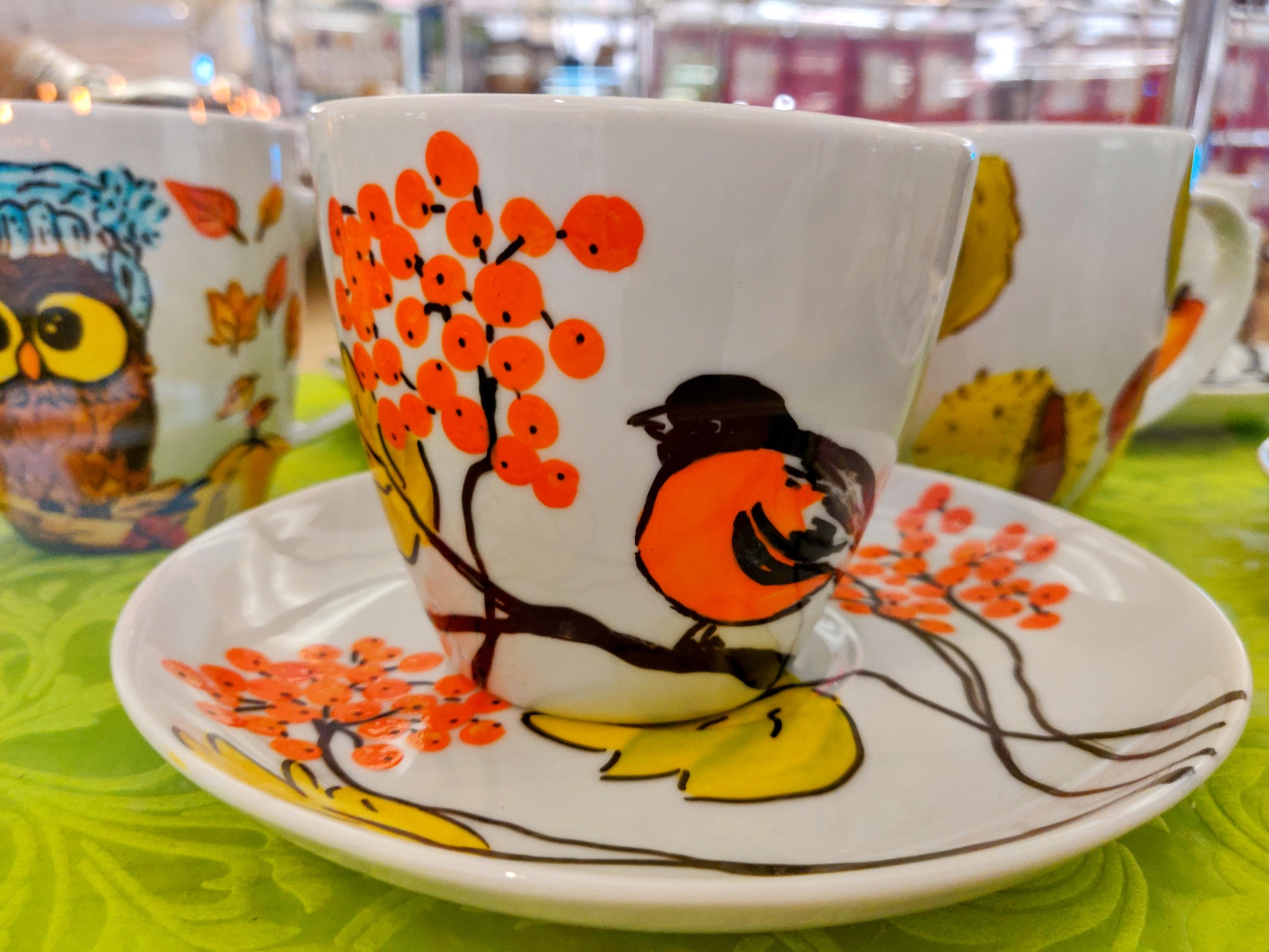 Biała filiżanka z motywem jarzębiny i ptaszka siedząca na dopasowanym talerzyku, obok widoczne inne filiżanki z motywami jesiennymi, całość na zielonym tle.