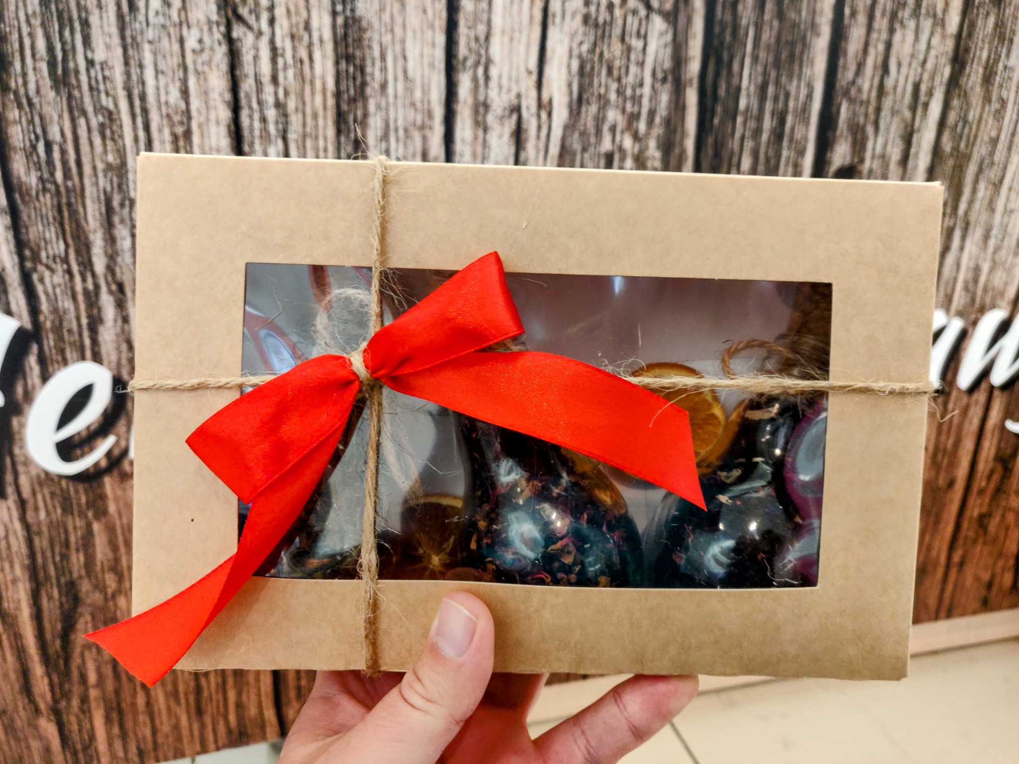 Ręka trzyma kartonowe pudełko prezentowe z okienkiem, przez które widać dekoracyjne świece i suszone pomarańcze, przewiązane sznurkiem jutowym i czerwoną wstążką na tle drewnianej ściany.