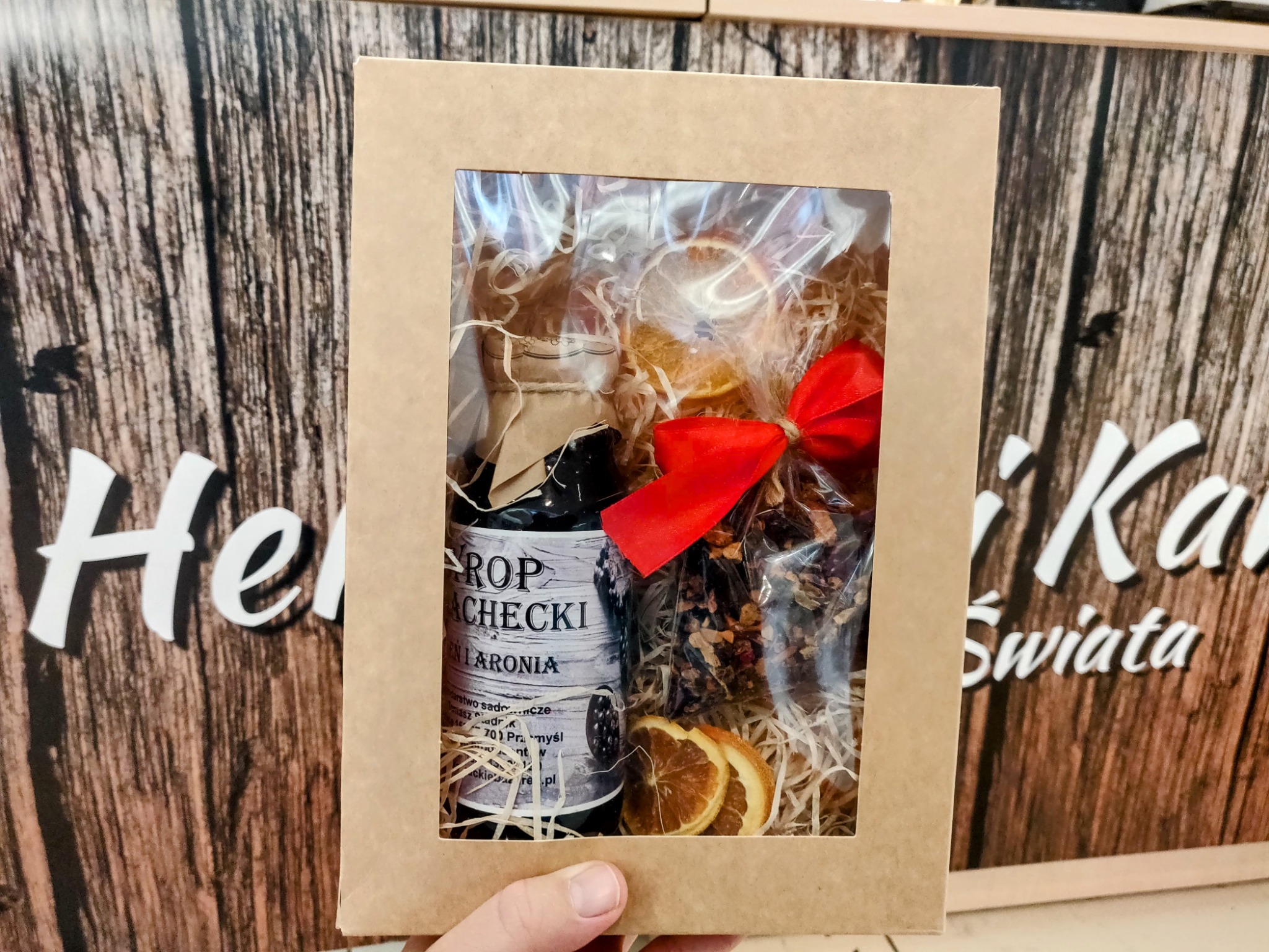 Prezent w kartonowym pudełku z okienkiem, zawierający butelkę syropu z aronii, suszone plastry pomarańczy i mieszankę herbaty, przewiązany czerwoną wstążką, na tle kartonowego opakowania...