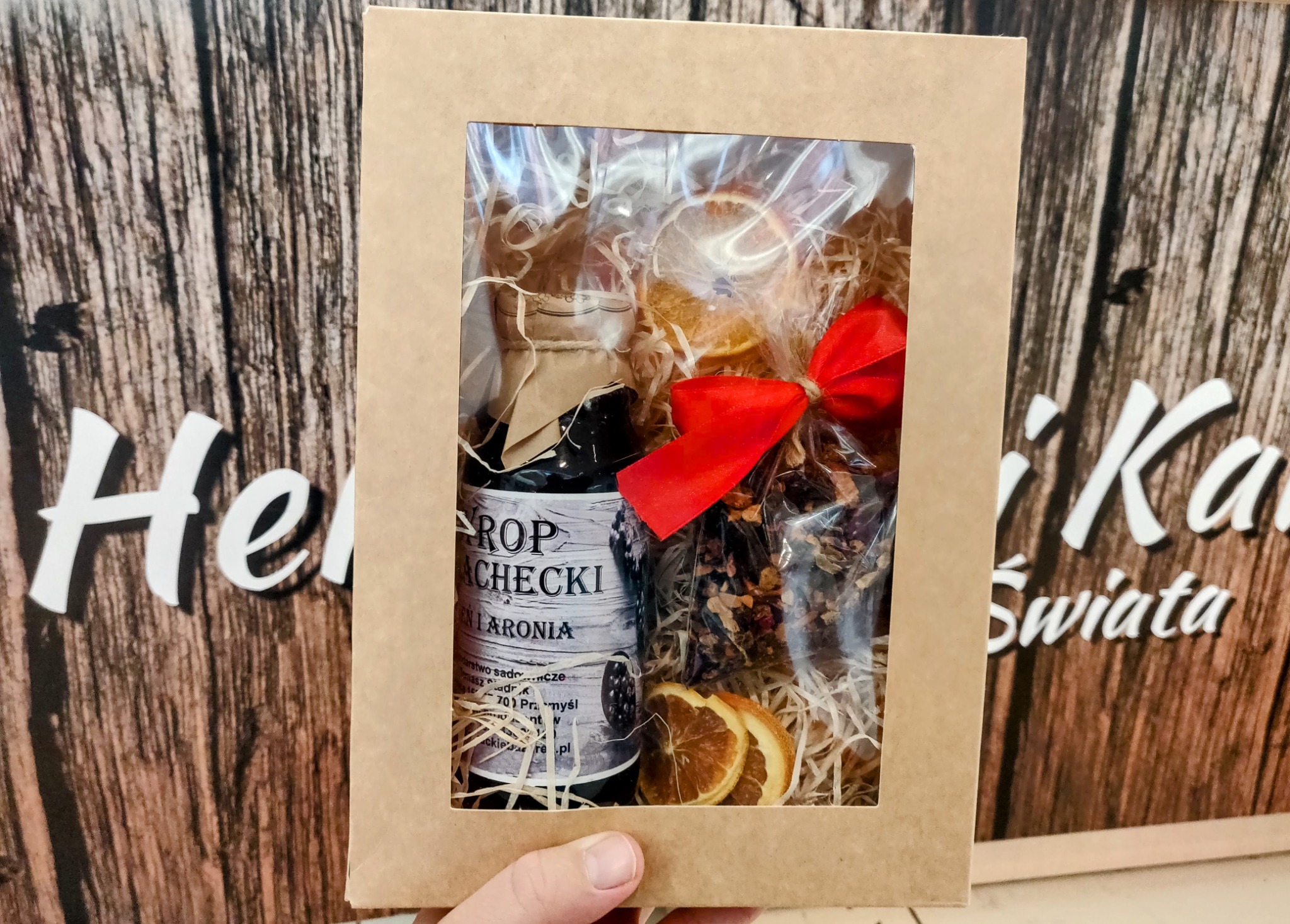 Ręcznie trzymany kartonowy prezent z okienkiem, wypełniony syropem z aronii w butelce, suszonymi pomarańczami i zapakowaną herbatą z czerwoną kokardką, na tle drewnopodobnej grafiki z napisem.