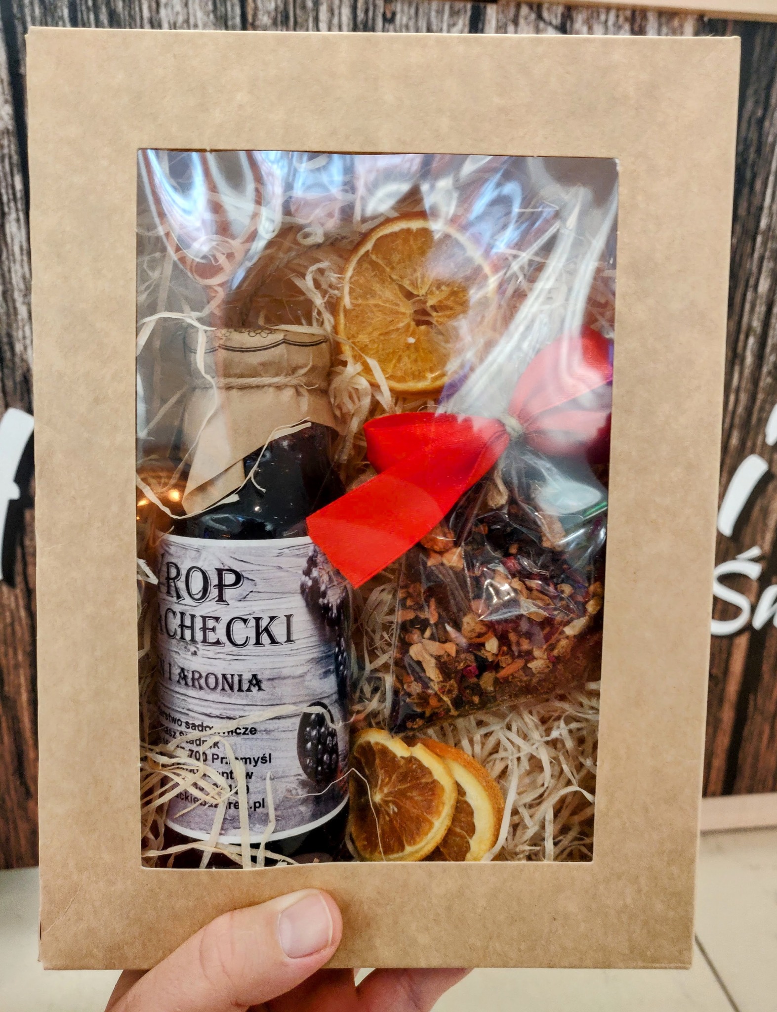 Ręcznie wykonany kosz prezentowy z dżemem z aronii, suszonymi pomarańczami i herbatą, zapakowany w tekturowe pudełko z okienkiem, przewiązany czerwoną wstążką.