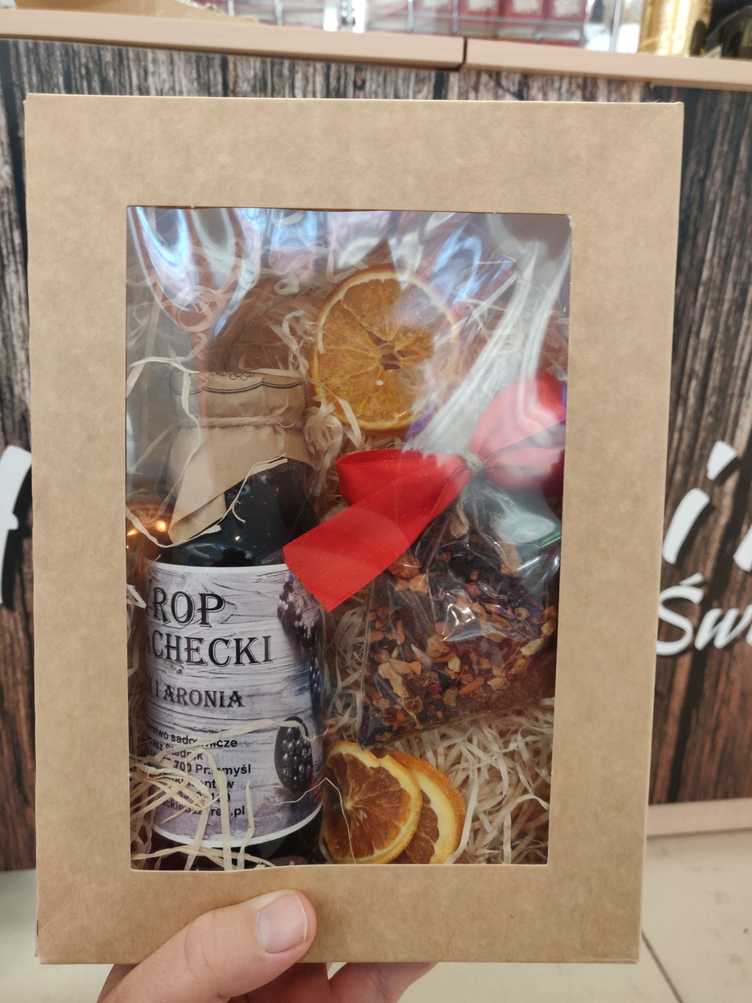 Prezent w kartonowym pudełku z okienkiem: syrop z aronii, suszone pomarańcze, czerwona wstążka i mieszanka herbat, wypełnione siankiem.