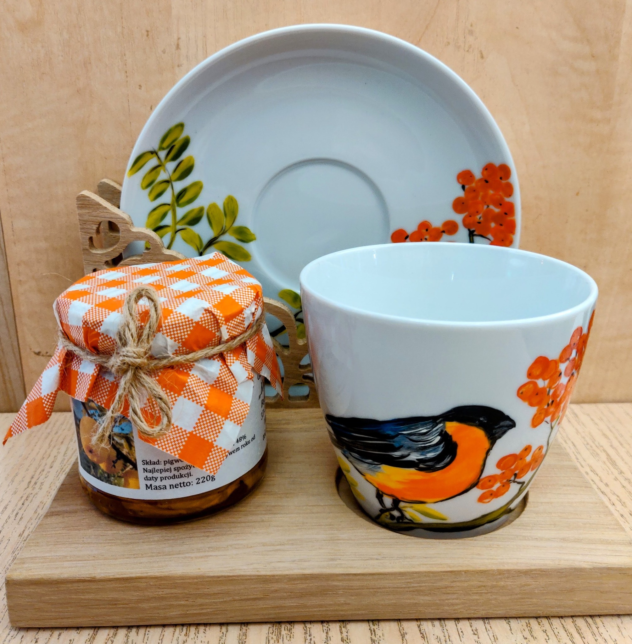 Słoik z dżemem pigwowym, nakryty pomarańczowo-białą kratką i sznurkiem, obok filiżanka z ręcznie malowanym ptakiem i jarzębiną oraz talerzyk z motywem roślinnym, na drewnianej desce.