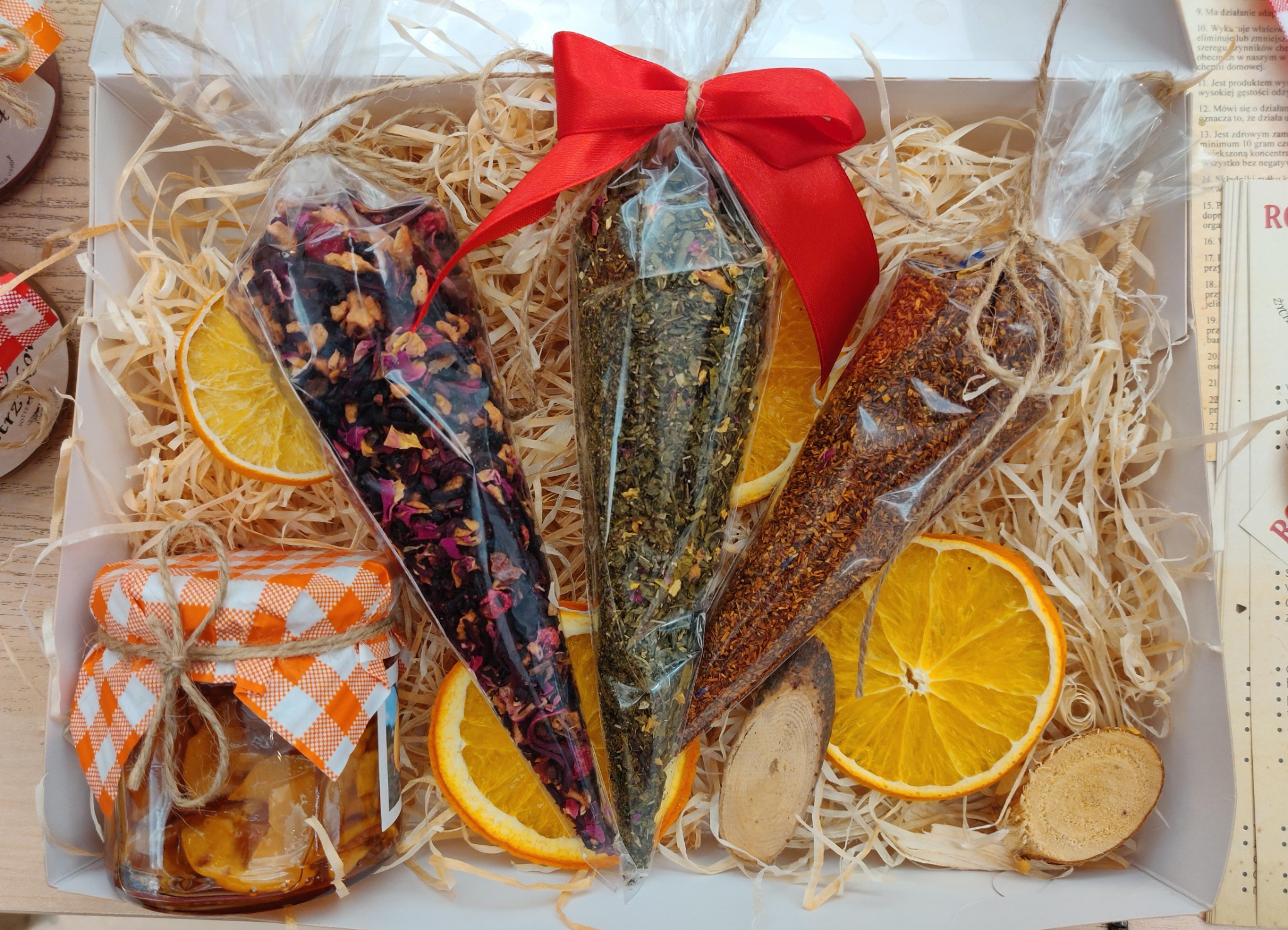 Kosz prezentowy z trzema przezroczystymi stożkowymi woreczkami z herbatą (różana, ziołowa, rooibos), plastrami pomarańczy, małym słoiczkiem dżemu z pomarańczową zakrętką w kratkę i drewnianymi...