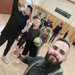 Grupa dzieci z zasłoniętymi oczami uczestniczy w zajęciach w sali gimnastycznej, prowadzonych przez animatorów, jeden z animatorów robi selfie.